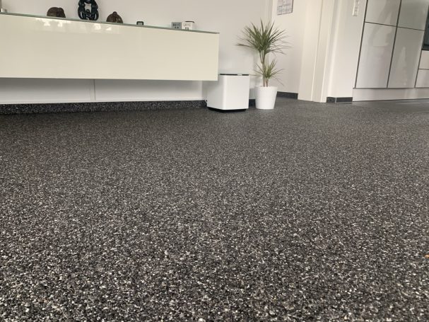 Nahaufnahme der dunklen Steinkörnung eines Steinteppichs vor einem weißen Sideboard.
